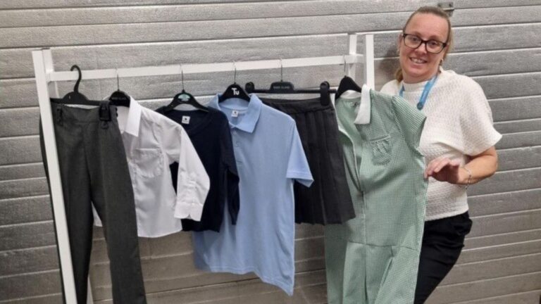 Rail of brand-new school uniform clothing including trousers, shirts, polo shirts, skirt and green gingham dress displayed on hangers, with a person standing beside the rack in a shop setting.