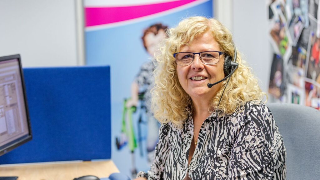 A Newlife Nurse wearing glasses and a headset sits at a desk in an office cubicle, facing the camera. A computer monitor, blue partition and colourful posters and photos are visible in the background.
