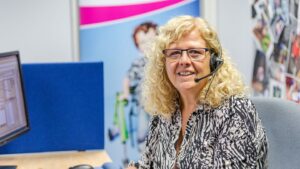 A Newlife Nurse wearing glasses and a headset sits at a desk in an office cubicle, facing the camera. A computer monitor, blue partition and colourful posters and photos are visible in the background.