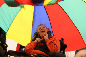 This picture shows a young child in a wheelchair sitting under a multicoloured sensory kite