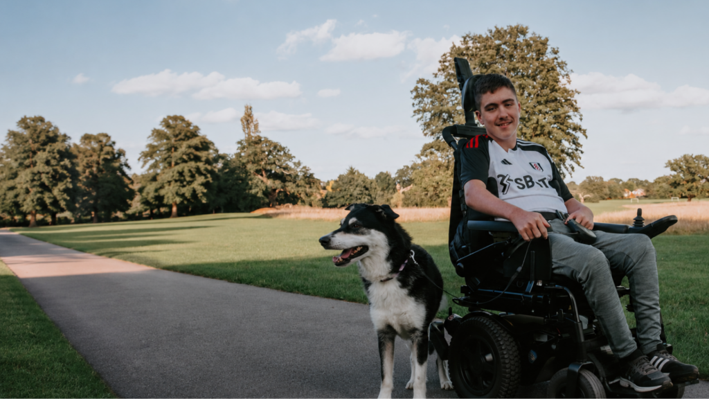This picture shows a teenage boy in a powered wheelchair provided by Newlife with his dog in a park setting