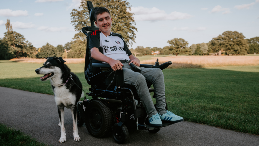 This picture shows a male teenager in a powered wheelchair provided by Newlife. In a park setting with his dog.