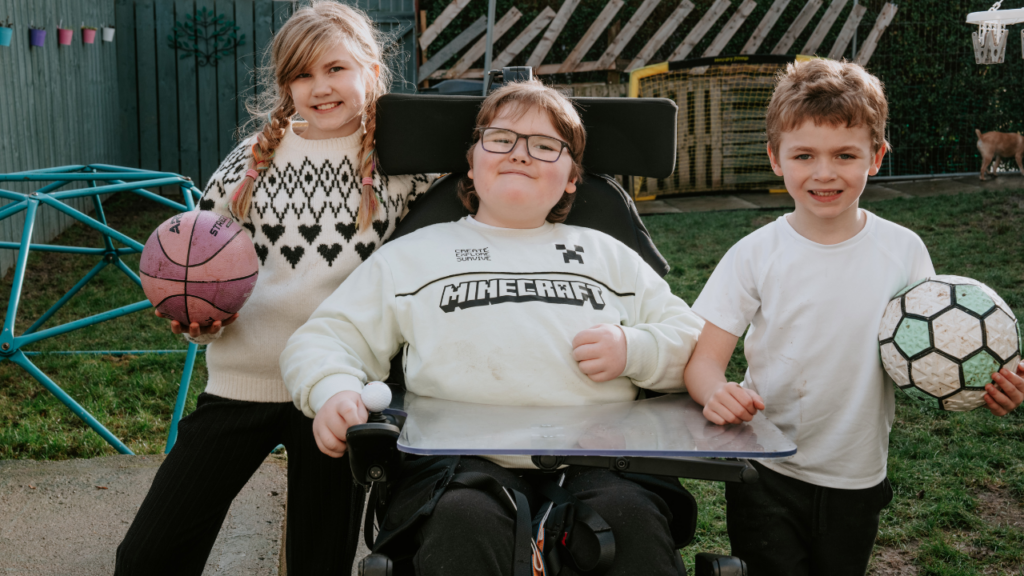 The picture shows a boy in a wheelchair with his sister and brother next to him.